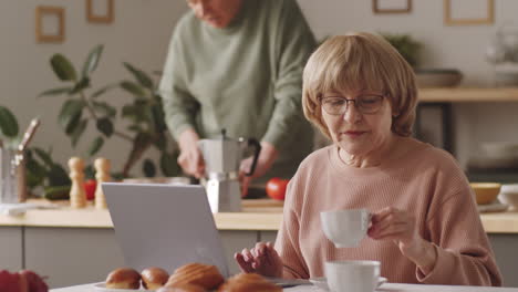 Ältere-Frau-Benutzt-Laptop,-Trinkt-Kaffee-Und-Spricht-Mit-Ehemann